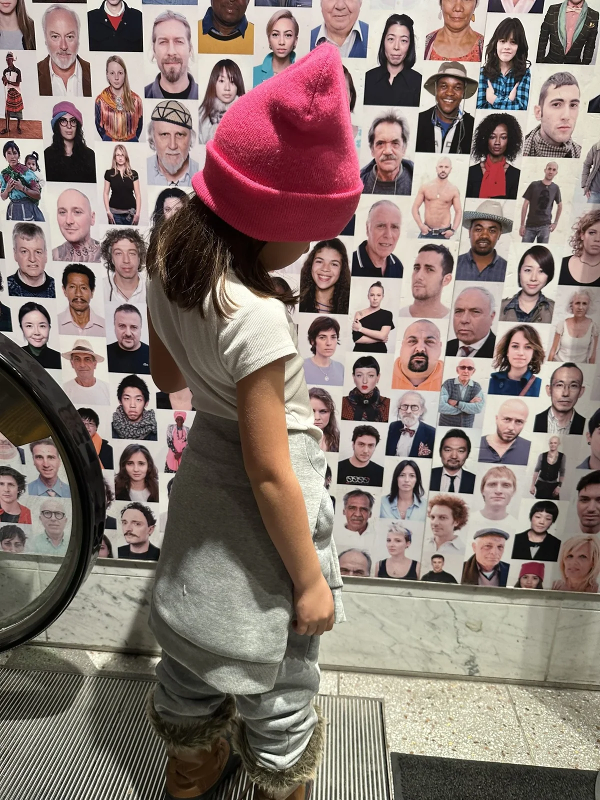 A child in a pink hat looking at a wall of portraits