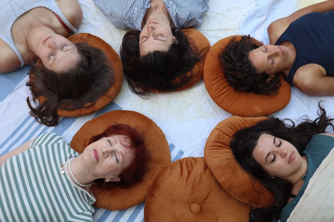 Six women participating in an intraspective meditation lying on their backs with eyes closed, resting on orange pillows, arranged in a circle.
