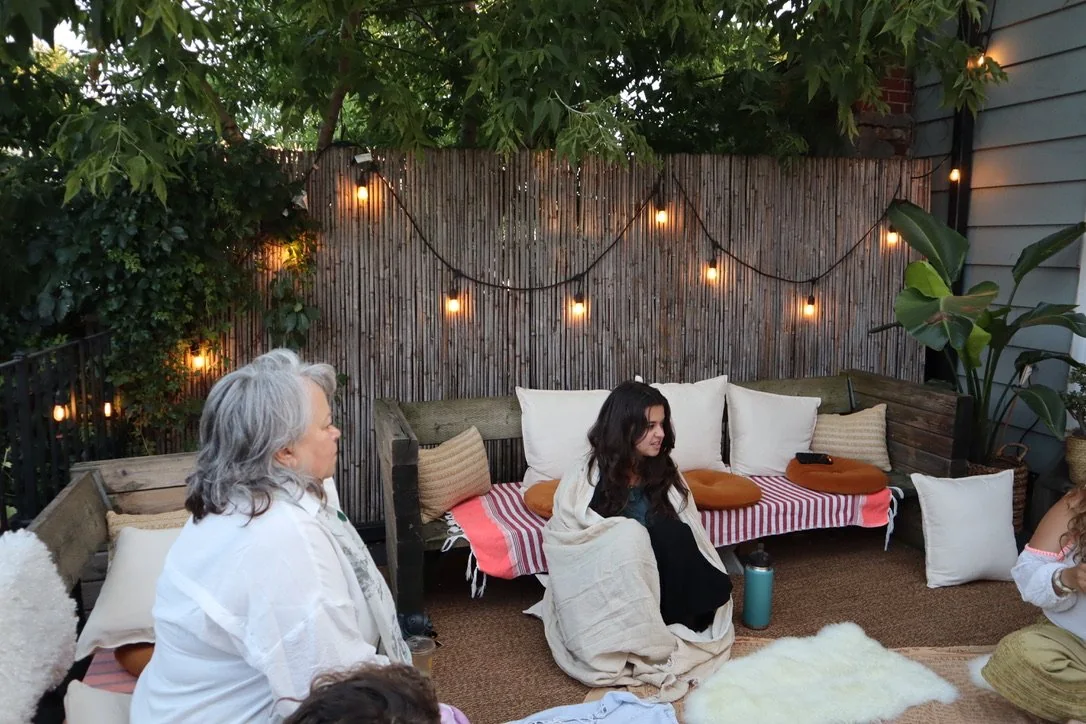 People sitting on cushions and blankets in a cozy outdoor space with string lights, potted plants, and a wooden fence. Taking part in a group psychotherapy session in the Intraspective Imaginarium at Mindful Maelstrom Wellness Clinic