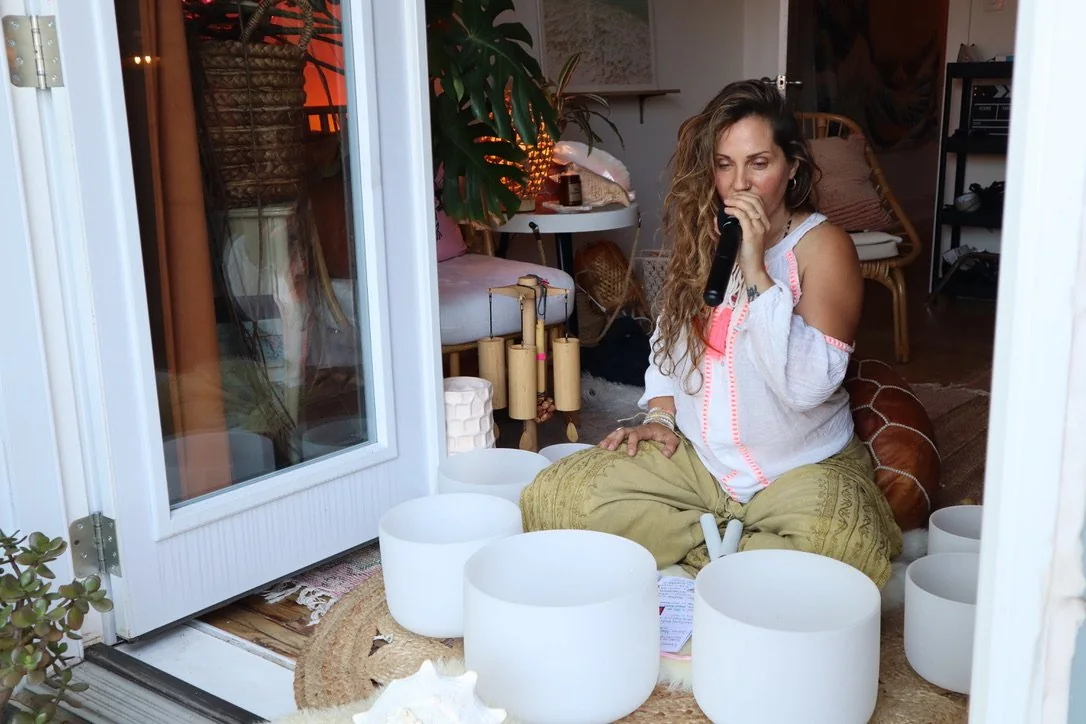 A woman with long curly hair sitting cross-legged on the floor, guiding intraspective meditation using a microphone, surrounded by white singing bowls inside a cozy room with plants and furniture.
